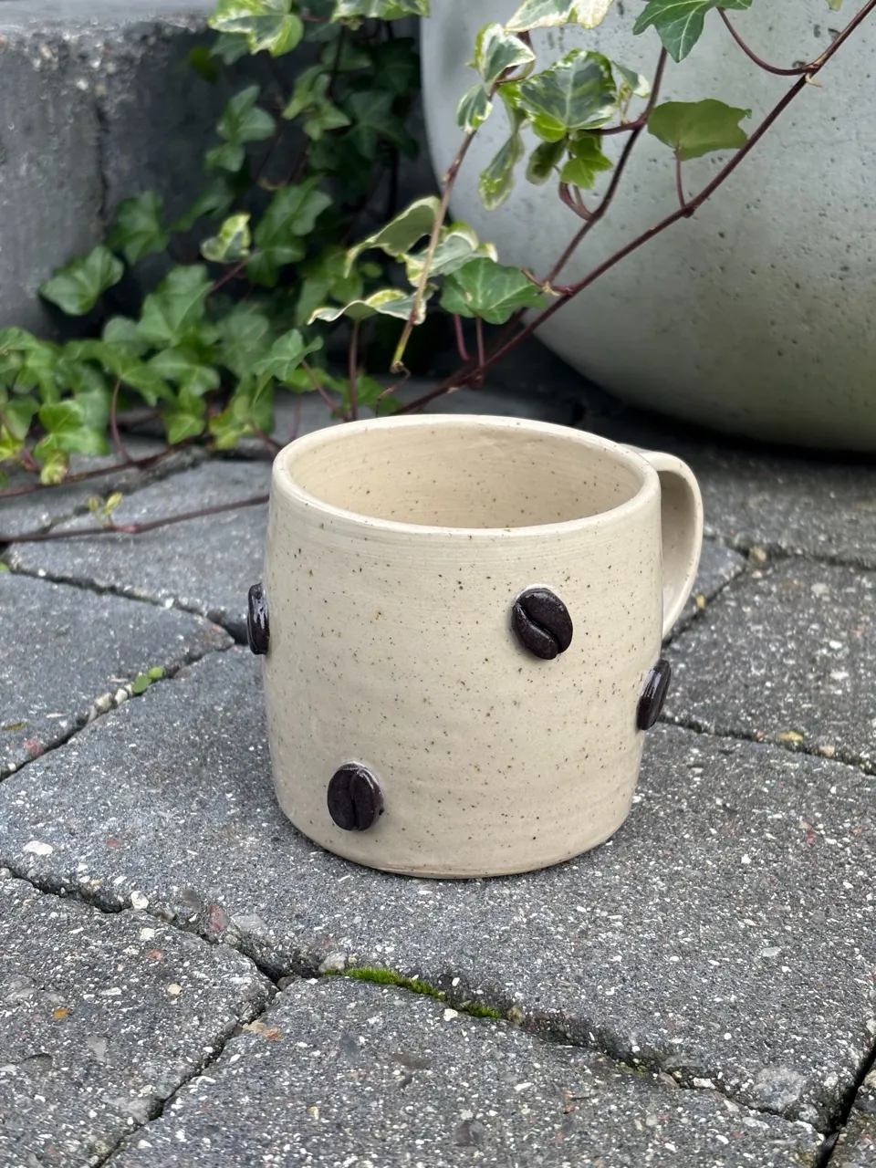 Ljus mugg med öra och mörkbruna kaffebönor pålagda runt sidan.