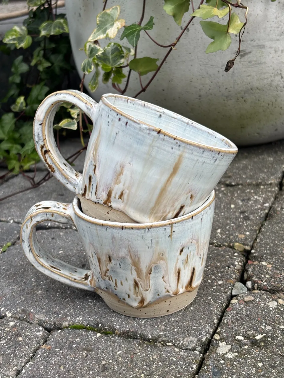 Två staplade koppar med öra och vit-brun rinnande glasyr över oglaserad fot.