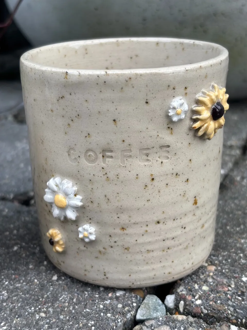 Sandfärgad mugg med ordet COFFEE instämplat och pålagda prästkragar och en solros.